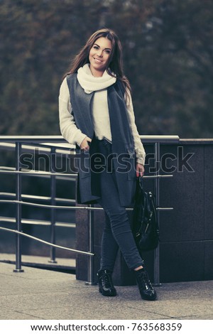 Happy young woman walking in city street. Stylish fashion model in long vest and white sweater outdoor