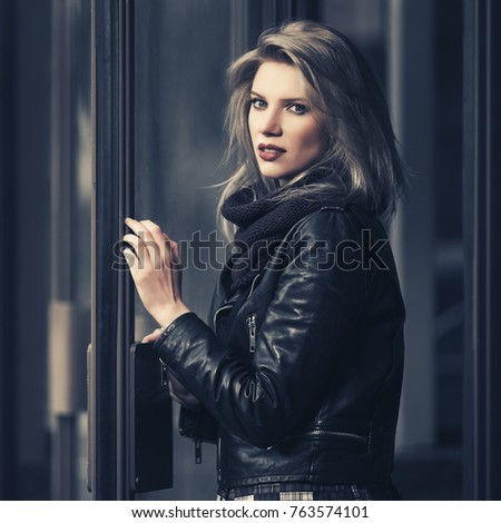 Young fashion blond woman at the mall door. Stylish female model in black leather jacket outdoor