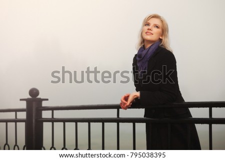 Young fashion woman in black coat leaning on handrail in a fog outdoor