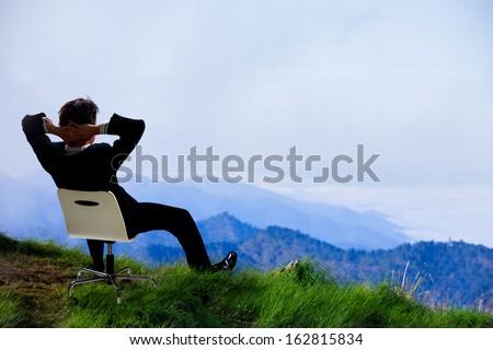 Young Businessman Who Sits On A Chair At The Top Of The Mountain And Looks Into The Sky
