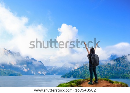 Young happy girl with backpack standing near big river with raised hands