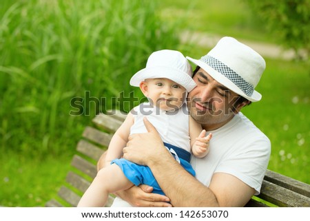 Father hugging his son with love. Sitting on the bench in the green park at summer time.