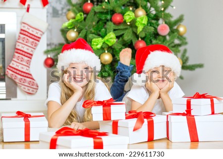 Happy brother and sister lying under Christmas Tree with gifts. Portrait of boy and girl smiling with Santa Hats at home, with decorated tree and chimney at background.