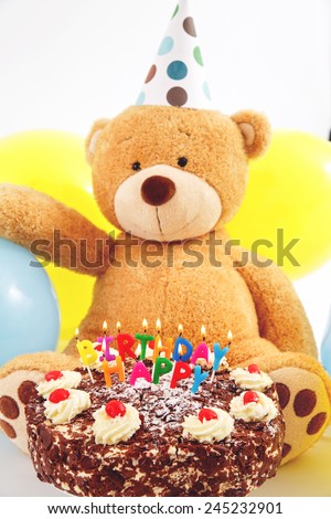 Teddy bear with birthday cap, balloons and cake with candles. Birthday greeting card. Happy birthday cake with burning candles.