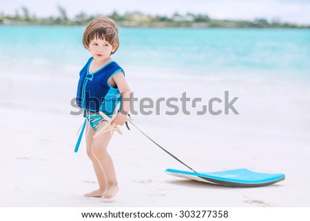 Cute baby with swimwear and life vest walking on the tropical beach with boogie board
