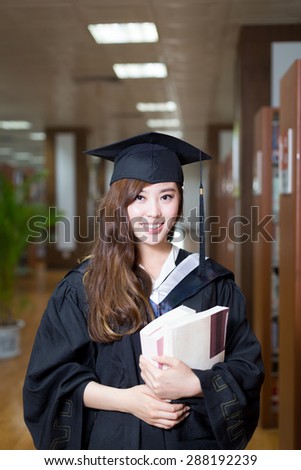 asian female student holding book and wearing academic dress in library