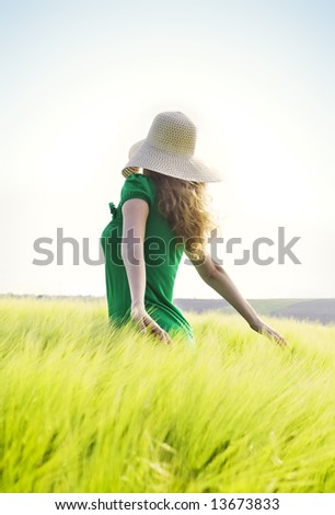 blond girl with big hat walking through a green wheat field against blue sky