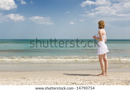 blond woman in white dress walking on the shore of a a Mediterranean beach looking at the horizon