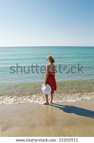 woman in red dress holding white hat standing on the beach and looking at the horizon