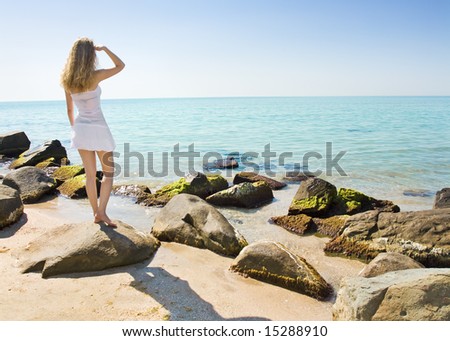 blond woman in white dress standing on the shore of a a Mediterranean beach looking at the horizon