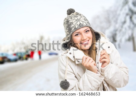 Beautiful Young Caucasian Teenage Girl Standing In Ski Center Park With Cars, Trees And Snowy Street Blurred In The Background. Winter Outdoors.