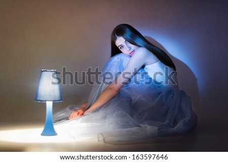Gorgeous young Caucasian ballerina sitting next to blue lamp. Dark background.