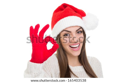 Excited young Caucasian brunette woman smiling wearing Santa Claus beanie hat and red gloves gesturing ok. Isolated on white background with available copy space.