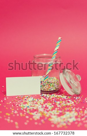 Transparent glass jar with colorful small sweets and candy and blank message next to it against pink background. Copy space available.