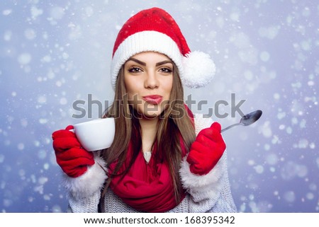 Closeup shot of cute happy Santa girl enjoying coffee in winter looking at camera making funny facial expression. Blurred snow on blue background. Christmas concept.