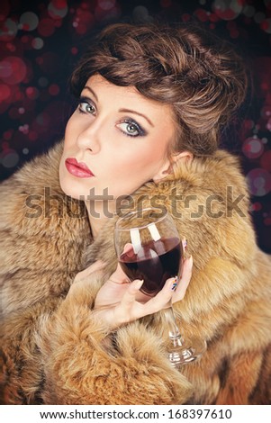 Elegant gorgeous young lady with fashionable hair style wearing brown fur coat celebrating Christmas drinking wine. Dark blurred background. Christmas, celebration and glamour concepts.