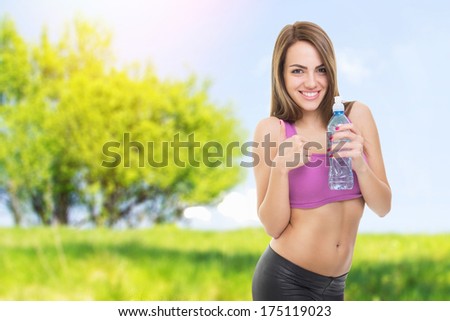 Fit happy young beautiful Caucasian woman outdoors fitness workout. Woman holding bottle of water and gesturing thumbs up. Fitness and healthy lifestyle concept.