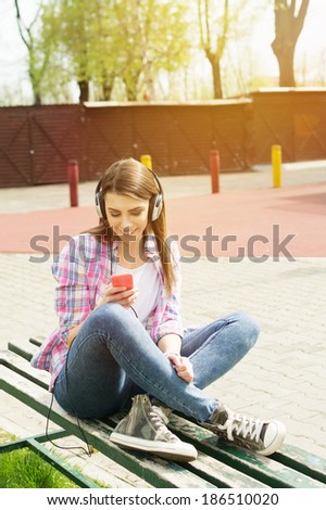 Happy beautiful young Caucasian brunette woman sitting on bench in park listening to music on headphones. Young people modern lifestyle concept.