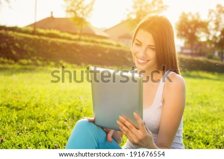 Happy young Caucasian brunette woman with tablet in park on sunny summer day sitting on grass, back lit, smiling, looking at tablet screen. Modern lifestyle and relaxation concepts.