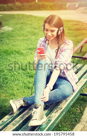 Cute hipster Caucasian teenage girl with smart phone and headphones listening music in park sitting on a bench. Modern young people lifestyle.