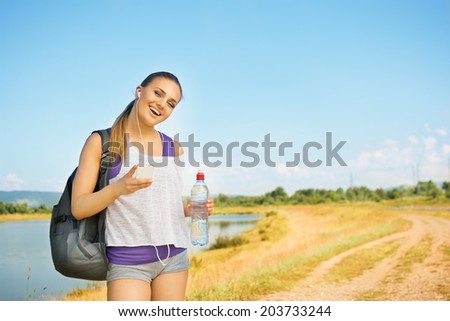 Beautiful young runner Caucasian blonde woman with smart phone outdoors in summer by the lake holding smart phone, bottle of water and backpack. Running, diet, healthy lifestyle concept.