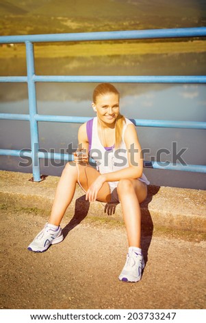 Young runner Caucasian blonde woman with smart phone and headphones making music playlist for jogging. Smiling looking at camera. Sunny summer day outdoors workout routine.