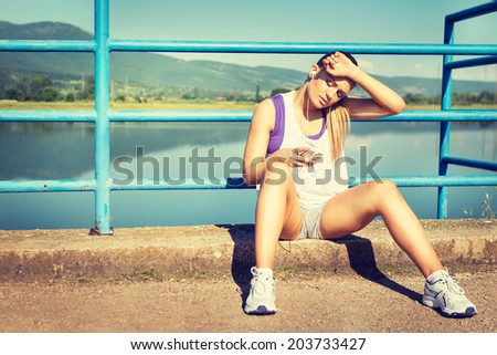 Tired young Caucasian blonde runner woman with smart phone and headphones sitting on the bridge resting from jogging wiping sweat listening to music. Summer outdoors sport.