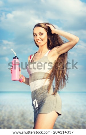 Beautiful fit young woman at the beach holding water bottle with long loose hair. Fitness Caucasian female jogging at the beach by the sea in summer. Sport, diet, workout, healthy lifestyle concept.