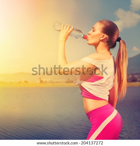 Young fit attractive blonde runner woman drinking water outdoors in summer by the lake on sunny summer day. Sunset color. Jogging teenage girl refreshing during workout outdoors.Instagram filter look.