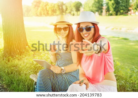 Two casual happy Caucasian teenage girls taking a selfie in park on sunny summer day. Two young women with hats and sunglasses enjoying outdoors in park taking a self portrait with smartphone.