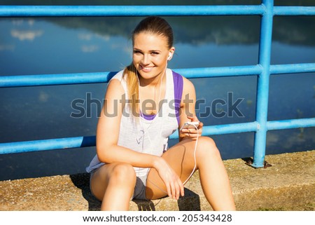 Pretty young runner woman with smart phone and headphones sitting by the lake on the bridge. Teenage fit blonde Caucasian girl in sportswear making a running playlist.