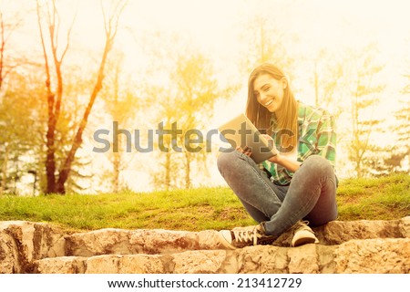 Young modern Caucasian teenage woman with tablet sitting in park laughing. Cute happy casual young woman with digital tablet. Warm sunset colors.