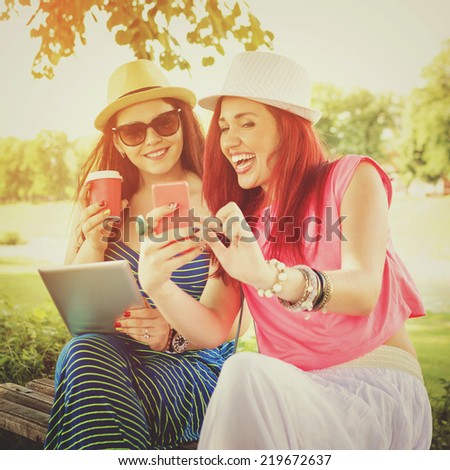 Two happy beautiful young Caucasian women with smart phone and digital tablet outdoors in park on sunny summer day.