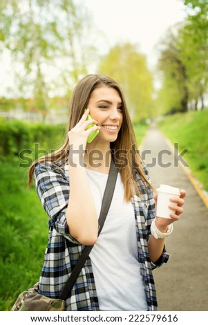 Beautiful young Caucasian woman with takeaway coffee speaking on the phone outdoors in park. Casual teenage girl with long blonde hair talking on smart phone smiling. No retouch.