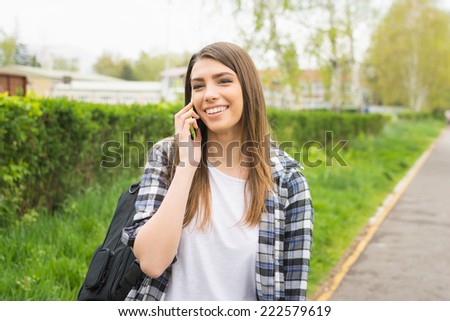 Beautiful young Caucasian woman with backpack speaking on the phone outdoors in park smiling. Cute teenage girl with checkered shirt and long blonde hair talking on smart phone. No retouch.