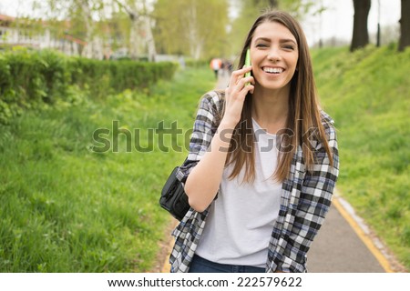 Pretty young Caucasian woman with backpack speaking on the phone outdoors in park laughing. Cute teenage girl with checkered shirt and long blonde hair talking on smart phone. No retouch.