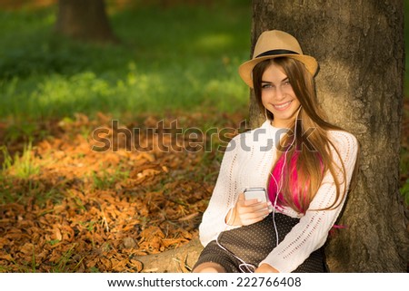 Beautiful hipster girl with long blonde and pink dyed hair and hat holding smartphone sitting in park in autumn smiling. Closeup of teenage girl using smart phone. No retouch.