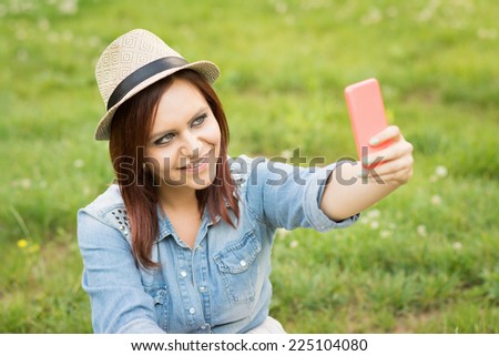 Young redhead Caucasian woman with hat taking a selfie outdoors. Cute girl taking a self portrait in park in summer wearing denim shirt and beige hat smiling and posing.