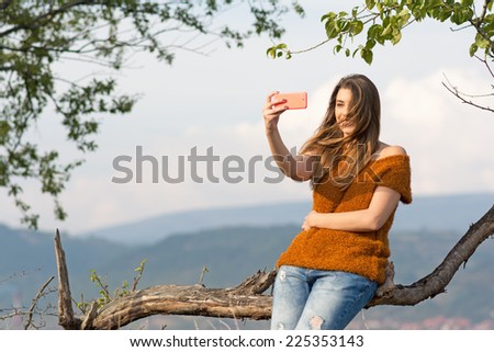 Young blonde Caucasian woman in park in autumn taking a selfie with smartphone. Beautiful teenage girl taking a self portrait outdoors in park on windy day. No retouch.