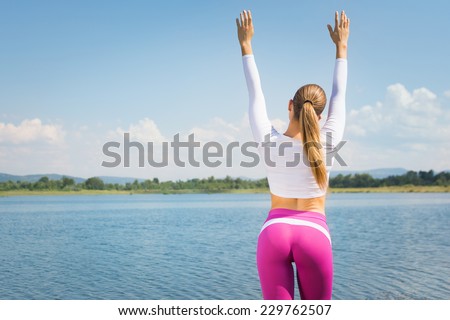 Rear view of young fitness woman with hands raised at the beach in summer. Attractive fit young woman in pink tights and ponytail standing by the sea in summer. Active lifestyle concept.