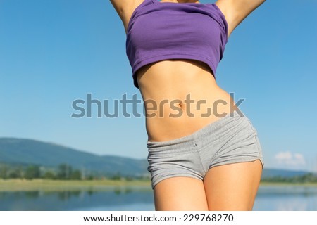 Closeup of young fit woman\'s belly outdoors in summer. Fitness girl in gray shorts and purple top outdoors at the beach. No retouch.