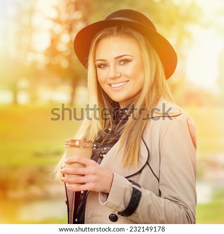 Closeup of beautiful young blonde Caucasian woman with fedora hat, beige coat and takeaway coffee outdoors in park on sunny autumn day. Square format image.