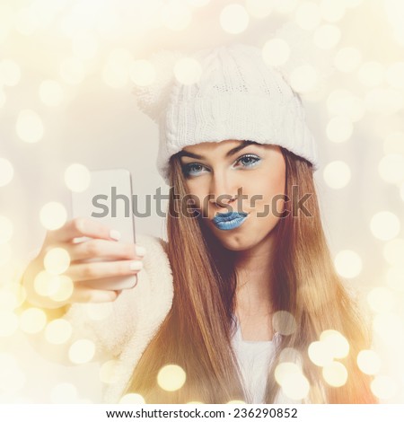 Closeup of cute teenage girl with beanie hat and blue lipstick taking a selfie with smart phone. Beautiful young woman with makeup photographing herself with smartphone. Square format image.