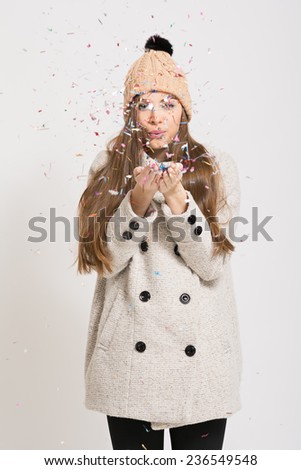 Trendy Caucasian teenage girl in beige coat and knitted beanie hat blowing confetti. Studio portrait of beautiful fashionable young woman with colorful confetti. No retouch.