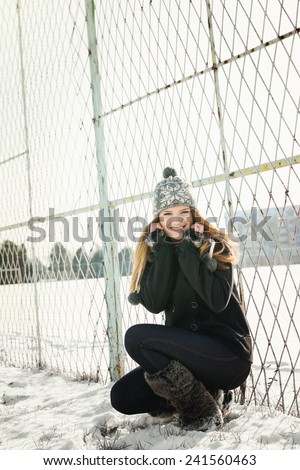 Cute blonde teenage girl in park in winter on snow wearing black coat, denim jeans, fuzzy boots and knitted gray hat smiling posing by fence. Vertical, no retouch, no filter.