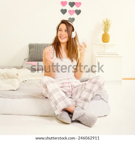 Young woman with headphones and smartphone in bed in the morning. Teenage girl in gray pink pajamas sitting in bed in modern contemporary decorated bedroom. Instagram filter, square format image.