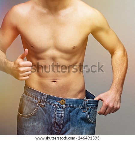 Shirtless muscular man wearing oversize jeans showing weight loss after diet. Closeup studio shot of unrecognizable fit young man showing thumbs up wearing big jeans. Square format, filter applied.