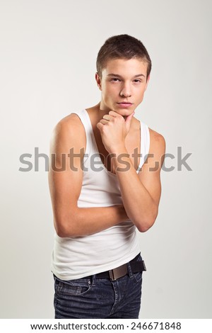 Handsome teenage male fashion model in white tank top and jeans. Closeup of skinny guy wearing white tank top and dark denim jeans with serious facial expression. No retouch.
