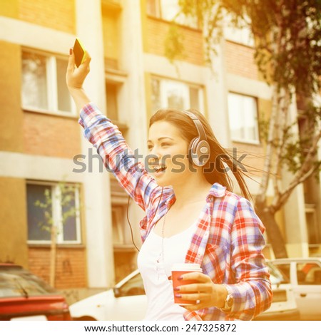 Cute happy teenage brunette girl with headphones, smartphone and takeaway coffee outdoors in urban environment with hand raised singing. Square format, warm colors, instagram look filter.