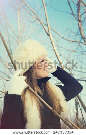 Gorgeous young Caucasian blonde woman with white fur hat, black coat and white gloves in park in winter. Matte filter, vertical, retouched.